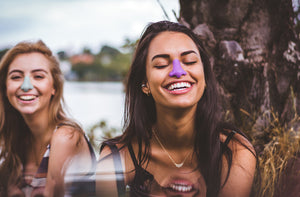 Two girls, sitting outside wearing colorful reef safe sunscreen on their noses.